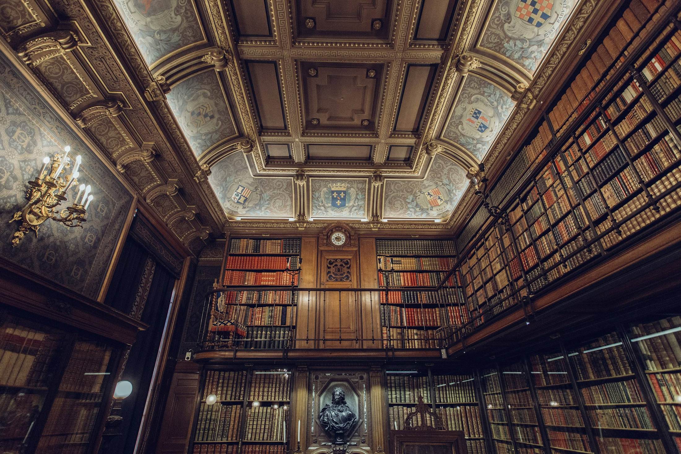 A panelled two-storey library with galleried bookcases and a painted ceiling, lit low.