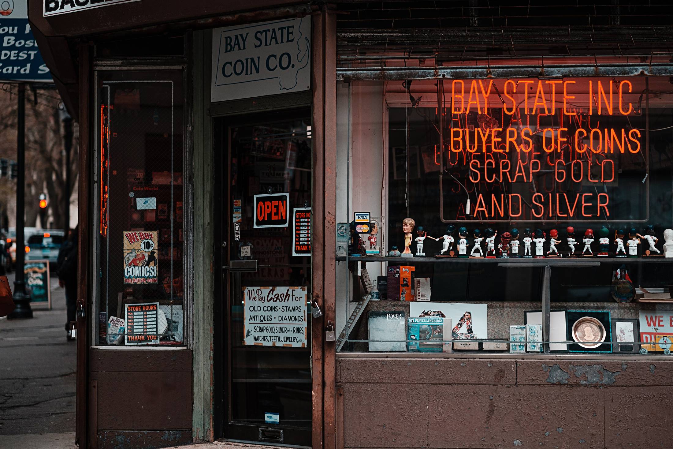 The window and doorway of an old independent shop on a quiet street, goods stacked behind the glass.