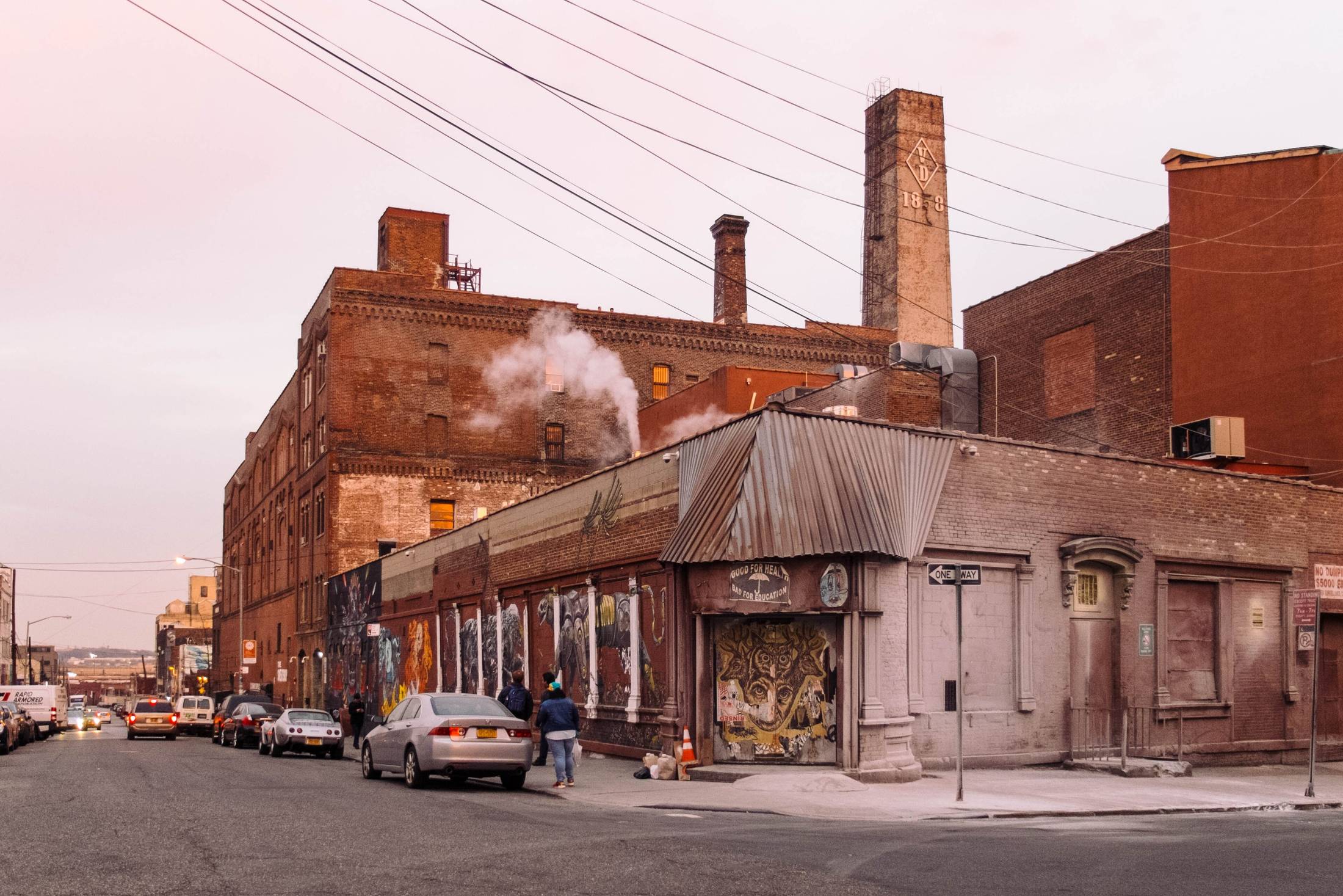 Brick industrial building on a corner lot at dusk, smokestack behind, wet street.