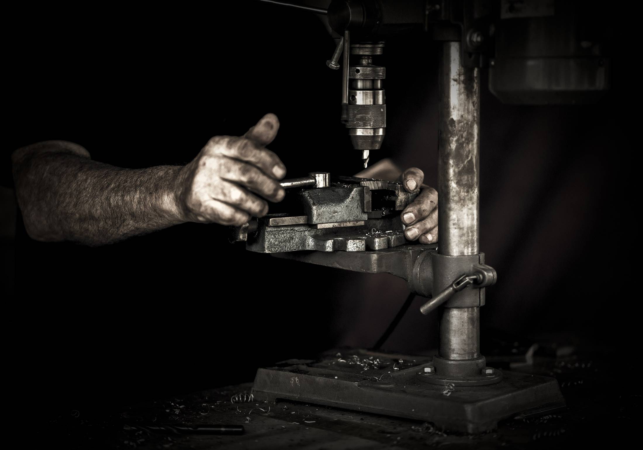 Two hands setting a part into the vice of a drill press, lit from one side against a dark shop.