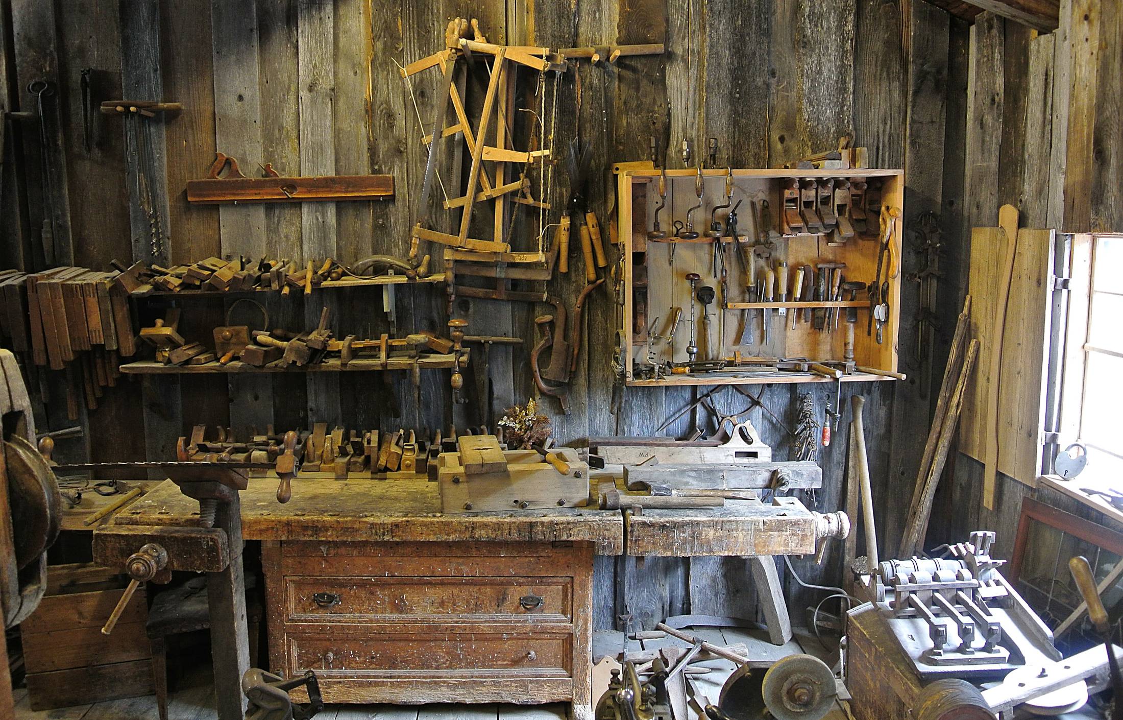 A timber workshop interior with a worn bench, hand tools racked on the wall and daylight from the right.