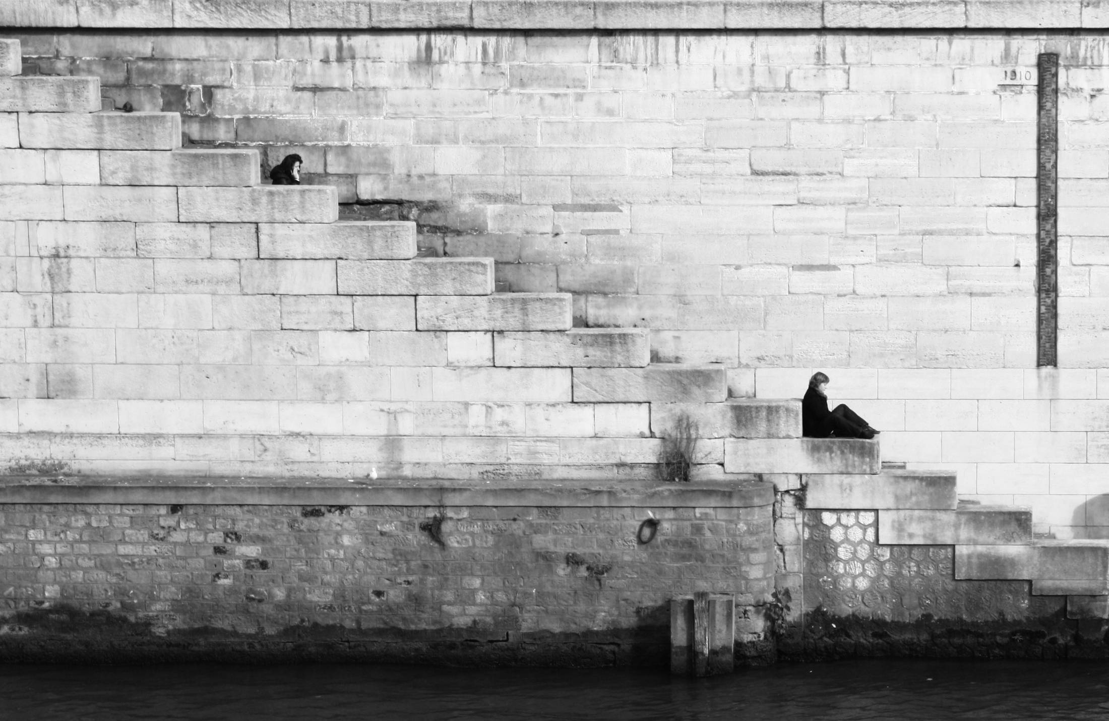 A monumental stepped stone seawall descending to still water, two figures small against it.